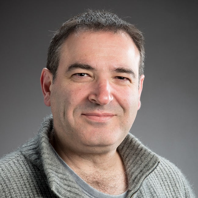 Portrait Photo of John Psathas wearing brown sweater and grey t-shirt.